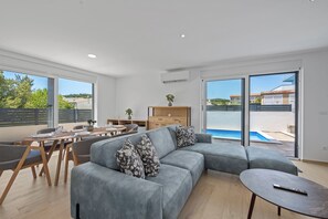 Ein modernes Wohnzimmer mit einem grauen Ecksofa, einem Esstisch mit Stühlen, einer kleinen Holzkommode und Blick auf einen Swimmingpool durch große Fenster.