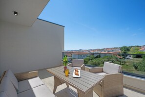 Ein Balkon mit Korbmöbeln, einem Tisch mit einem Croissant und einem Getränk, und eine Aussicht auf Gebäude in der Ferne.