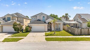 A residential street with houses featuring front garages, a central house with a prominent arched entrance, and a small palm tree. - Golden Light Retreat | 5BR Davenport w/ Pool (Davenport)