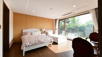Un dormitorio con una cama grande, una cama más pequeña, una silla y una ventana con vista a follaje.