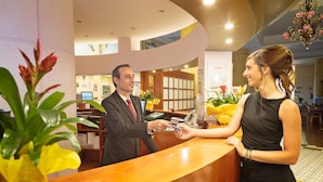 Zona de recepción de un hotel con un mostrador de madera, un hombre con traje entregando una tarjeta de crédito a una mujer con vestido negro.