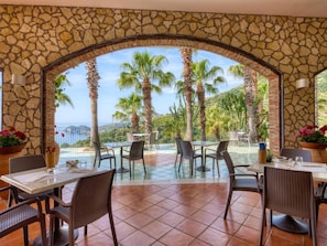 An outdoor dining area with stone walls, tables, and chairs, overlooking a scenic view with palm trees and a body of water. - Olimpo & Le Terazze (Letojanni)