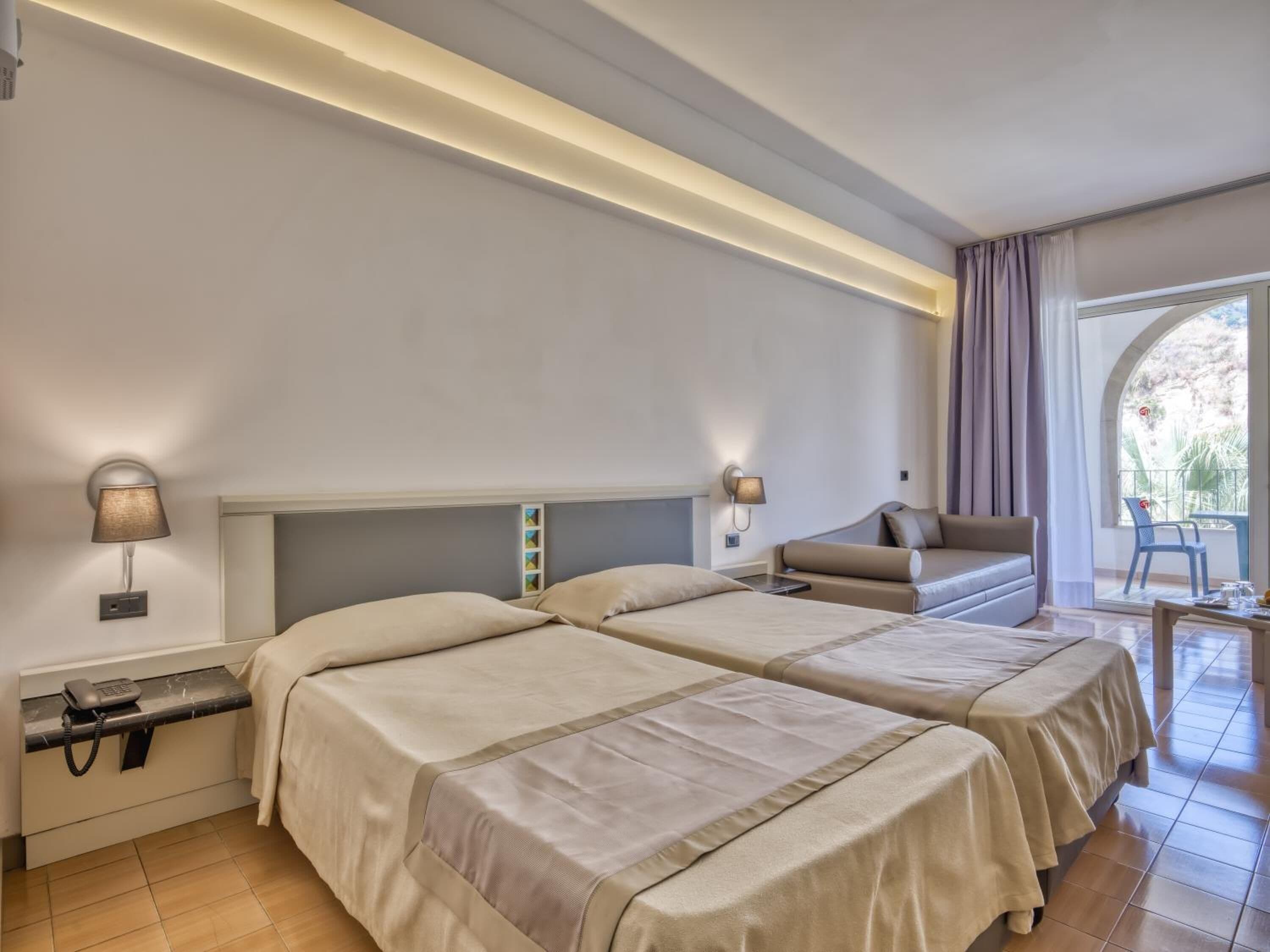 Quarto de hotel com duas camas, um sofá, uma mesinha e vista para o exterior.