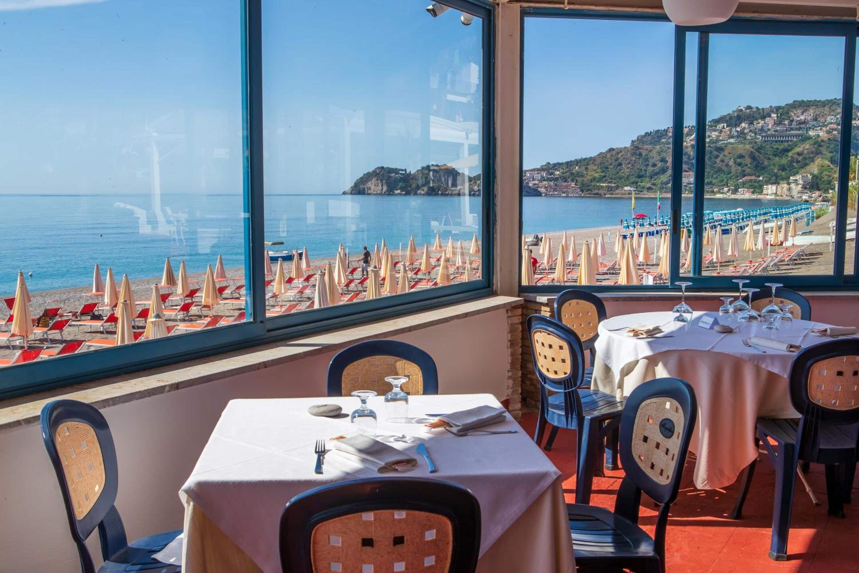 Restaurante com mesas e cadeiras dispostas para refeições, com vista para uma praia com guarda-sóis e espreguiçadeiras.