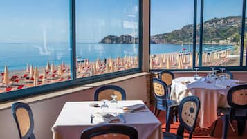 Restaurante com mesas e cadeiras dispostas para refeições, com vista para uma praia com guarda-sóis e espreguiçadeiras.