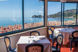 Un restaurante con mesas y sillas dispuestas para comer, con vista a una playa con sombrillas y sillones de descanso.