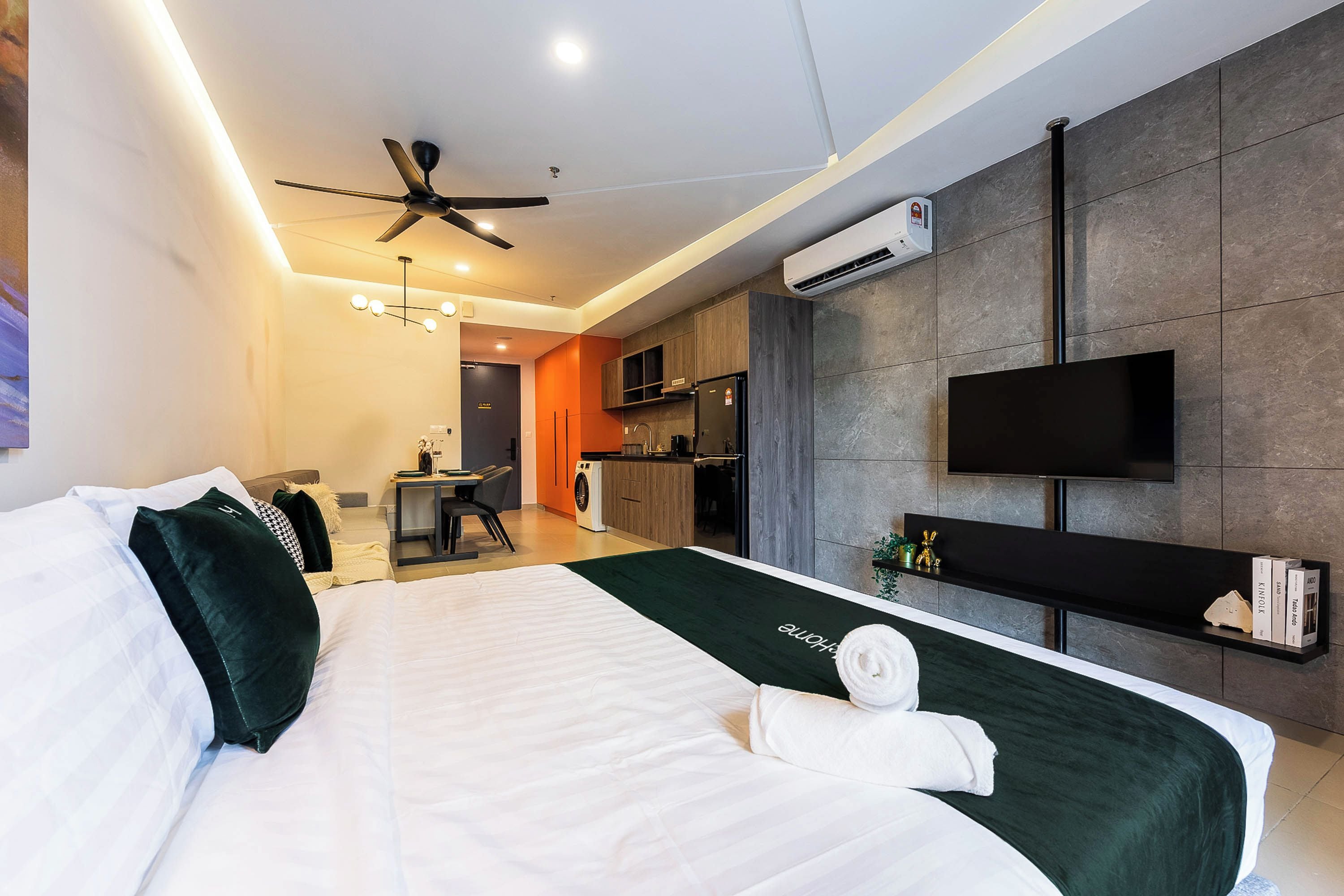 A modern hotel room with a bed, a desk, a kitchenette, and a TV mounted on the wall.
