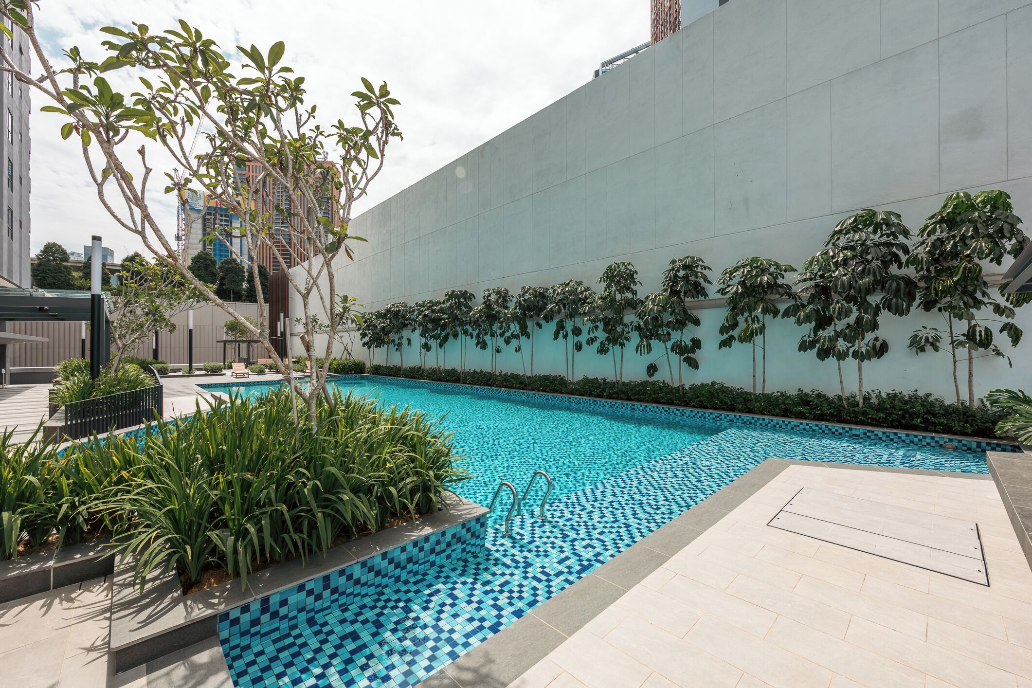 Una piscina con borde de azulejos, rodeada de follaje y una fachada de edificio moderna.