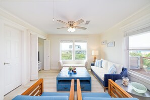 A bright living room with a ceiling fan, a sofa, a coffee table, and a view of the outdoors through large windows.