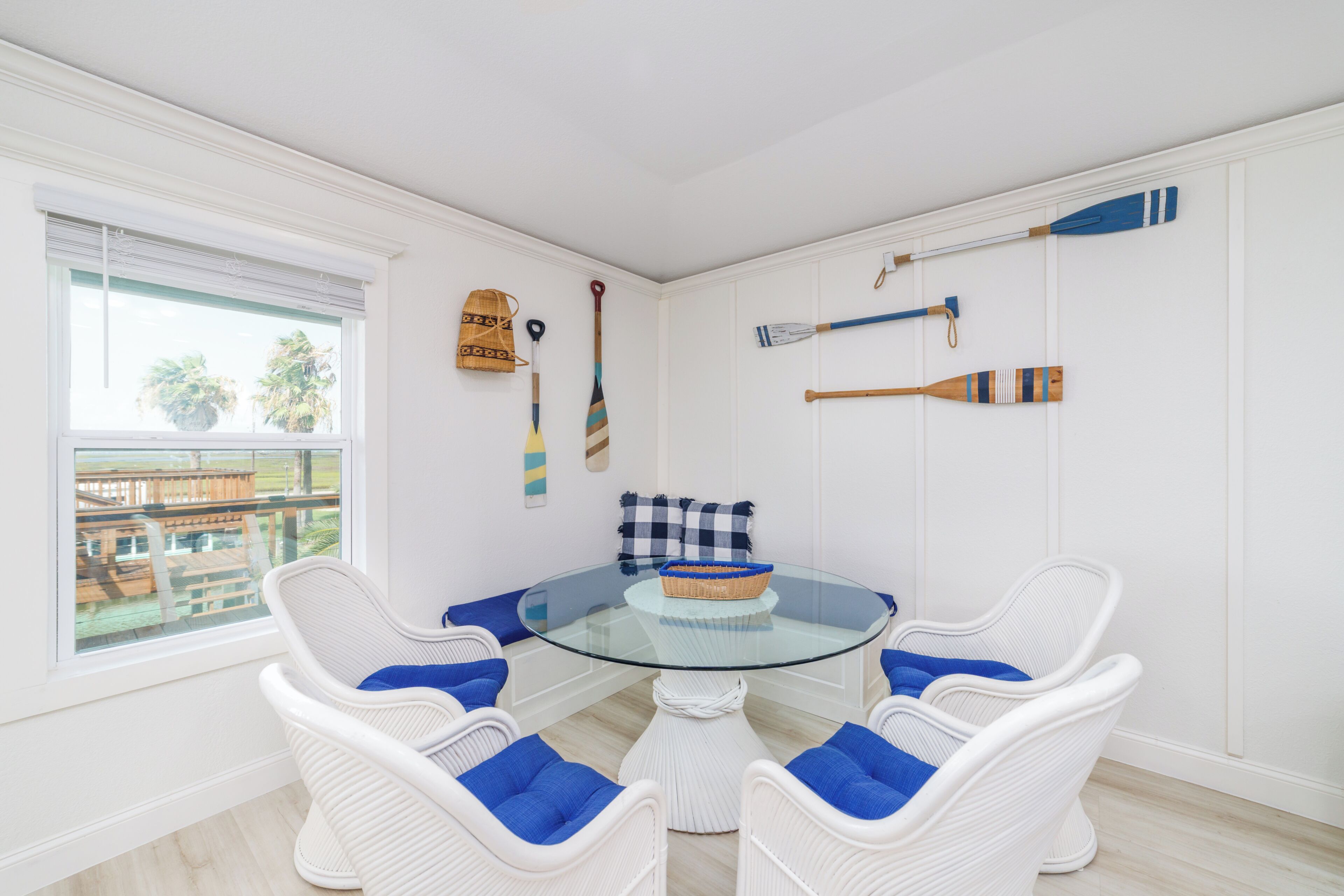 A room with a glass-top table surrounded by white wicker chairs with blue cushions, a checkered pillow, and a wicker basket.