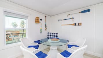 A room with a glass-top table surrounded by white wicker chairs with blue cushions, a checkered pillow, and a wicker basket.