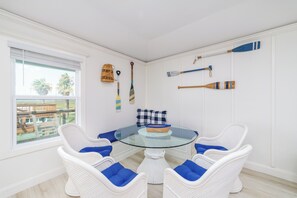 A room with a glass-top table surrounded by white wicker chairs with blue cushions, a checkered pillow, and a wicker basket.