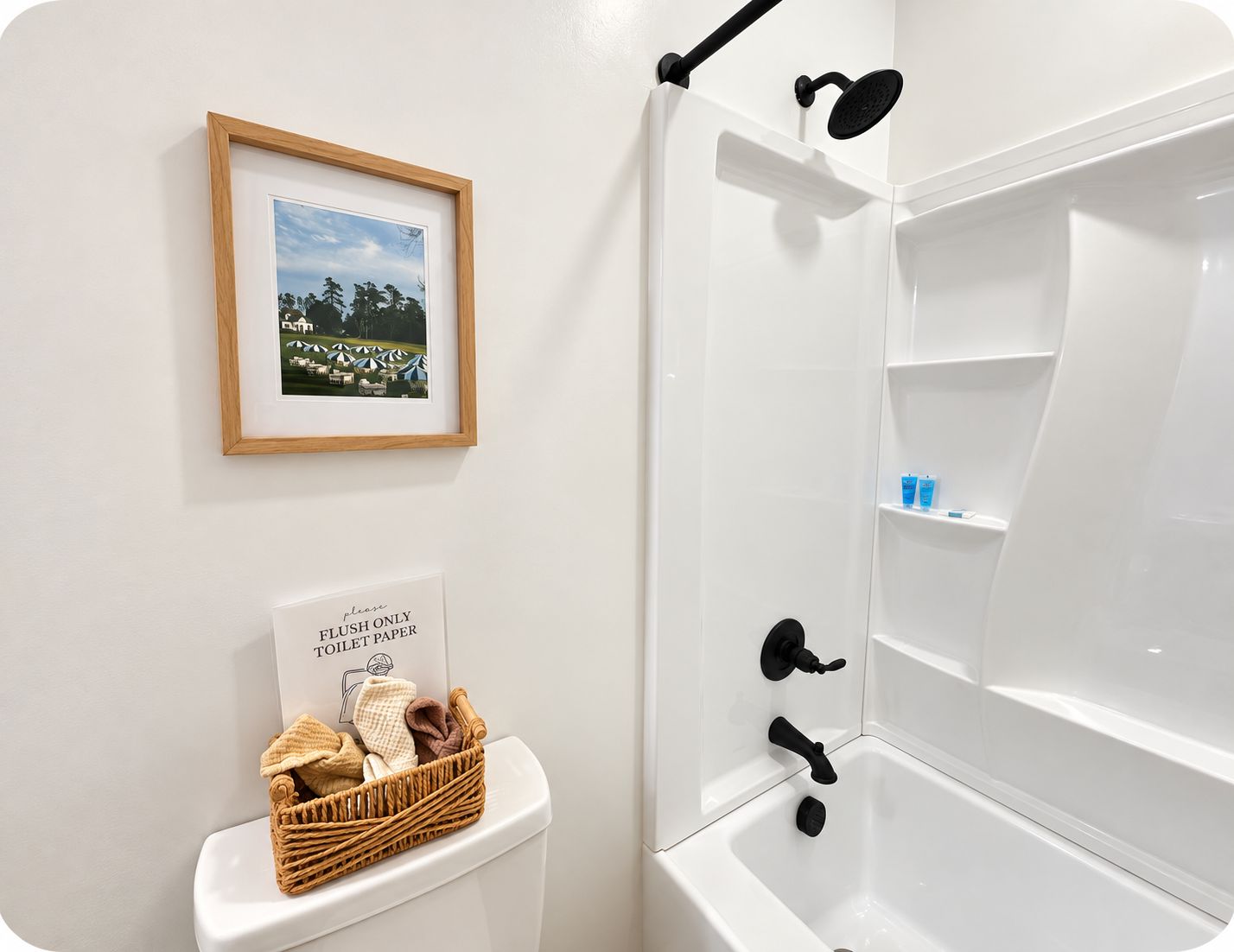 A bathroom with a framed picture on the wall, a sign instructing to flush only toilet paper, and a basket of towels on the toilet.