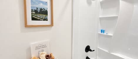 A bathroom with a framed picture on the wall, a sign instructing to flush only toilet paper, and a basket of towels on the toilet.