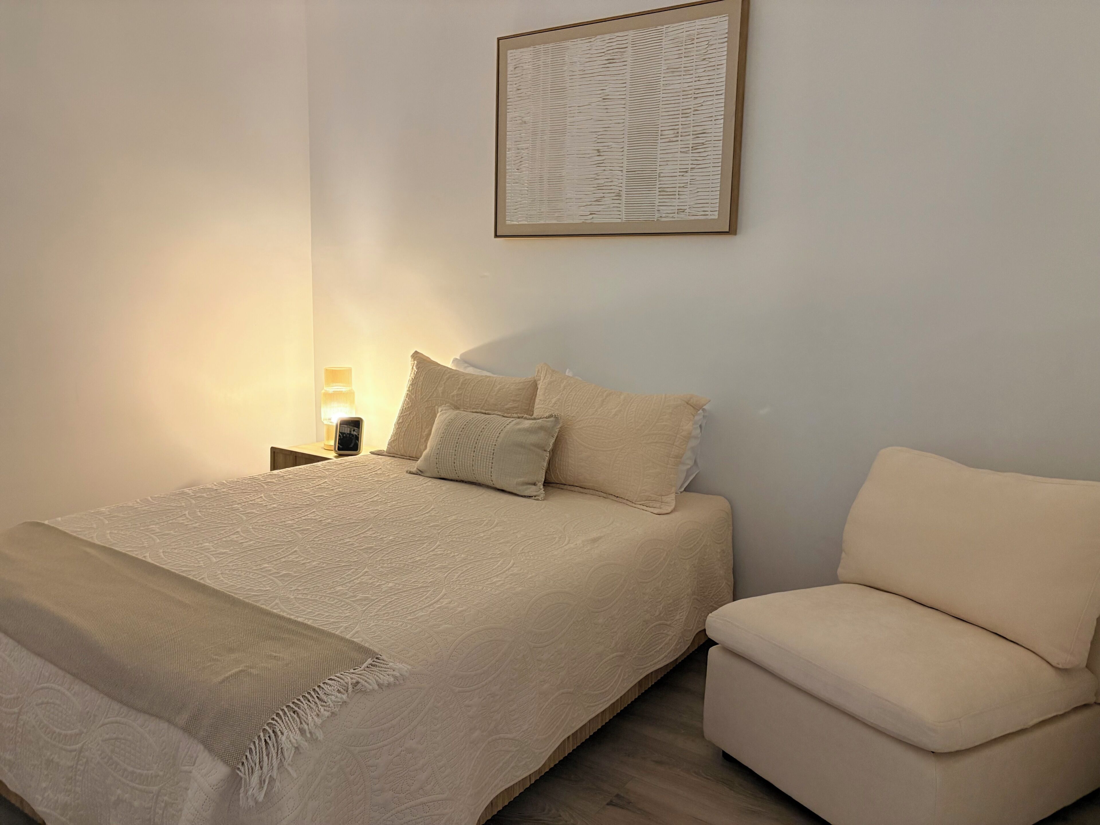 Une chambre à coucher avec un lit, une table de chevet, une lampe et une œuvre d’art encadrée accrochée au mur.