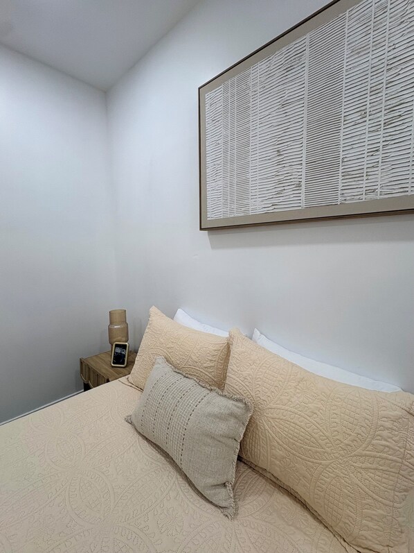 Une chambre à coucher avec un lit, des oreillers, une table de chevet, une lampe et une œuvre d’art encadrée accrochée au mur.