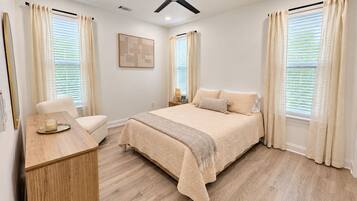 A bedroom with a bed, a bedside table, a chair, a ceiling fan, and windows with curtains.