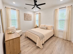Une chambre à coucher comprenant un lit, une table de chevet, une chaise, un ventilateur de plafond et des fenêtres avec des rideaux.