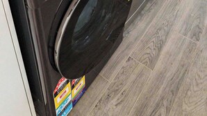 Eine kompakte Waschmaschine und Trockner-Kombination mit Bedienfeld, einer grünen Waschmittelflasche und einem Warnhinweis auf dem Boden.