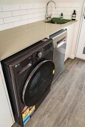 A compact washer and dryer unit with a control panel, a green detergent bottle, and a caution label on the floor. - Near city 2br house, walk to IGA, pub, restaurants/cafe, bus to city and beaches (Wembley)