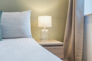 Ein ordentlich bezogenes Bett mit weißer Decke und Kissen, ein Nachttisch mit Lampe und ein teilweise gezogener Vorhang.