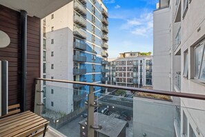 Ein Balkon mit einer hölzernen Bank, mit Blick auf ein Wohngebiet mit mehrstöckigen Gebäuden und einem klaren Himmel.