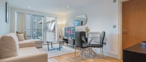 A modern living room with a sofa, a dining table with chairs, a TV, and a large window with curtains.