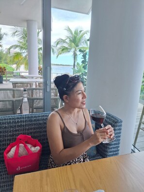 A person seated at a table with a drink, overlooking a beach and palm trees. - 2 bedrooms, 2 baths, luxury, most beautiful beach, Ocean view. 46 nights minimum (Nassau)