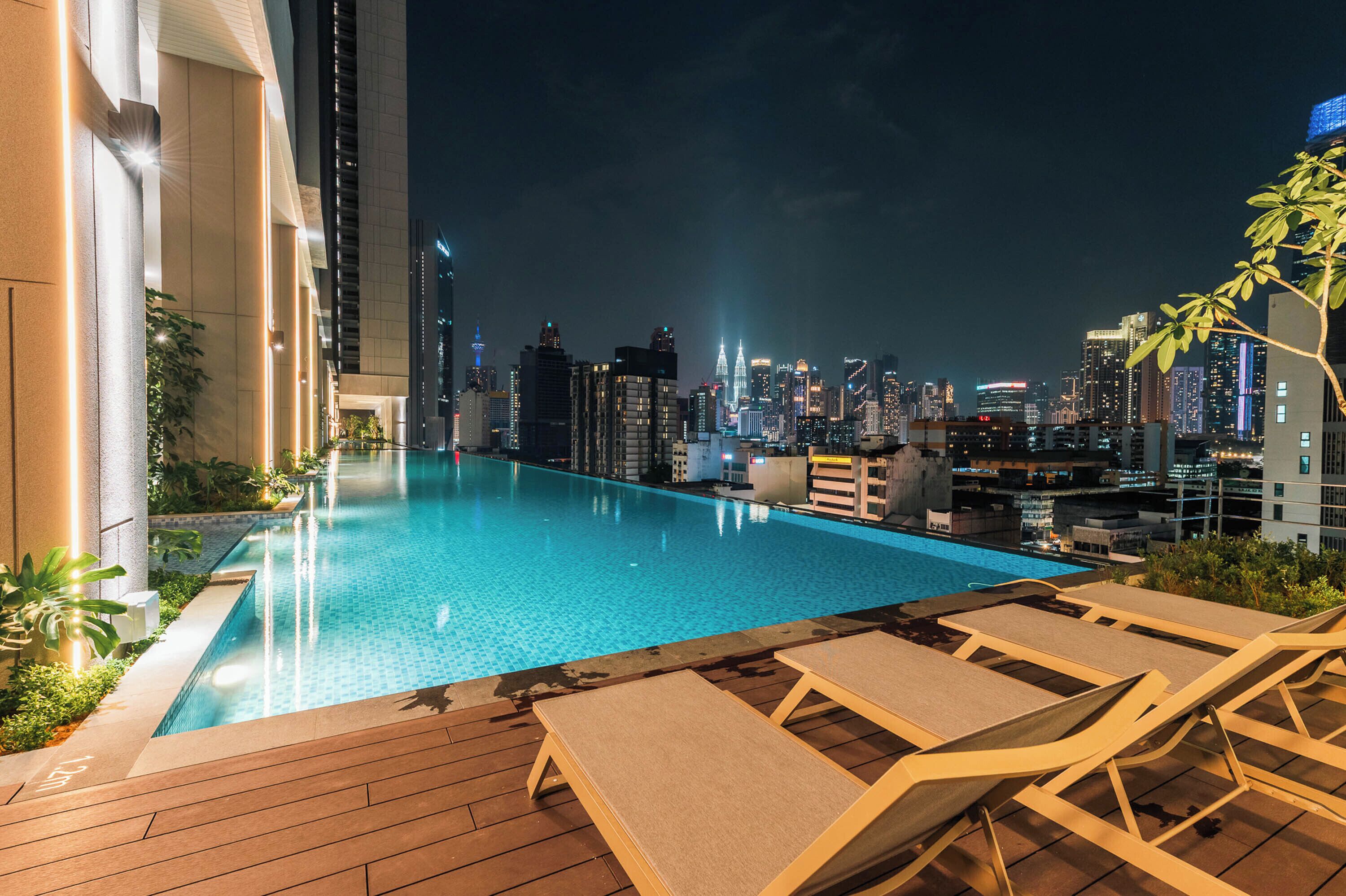 Une piscine sur le toit, agrémentée de chaises longues, et entourée d’une vue panoramique sur la ville illuminée la nuit.