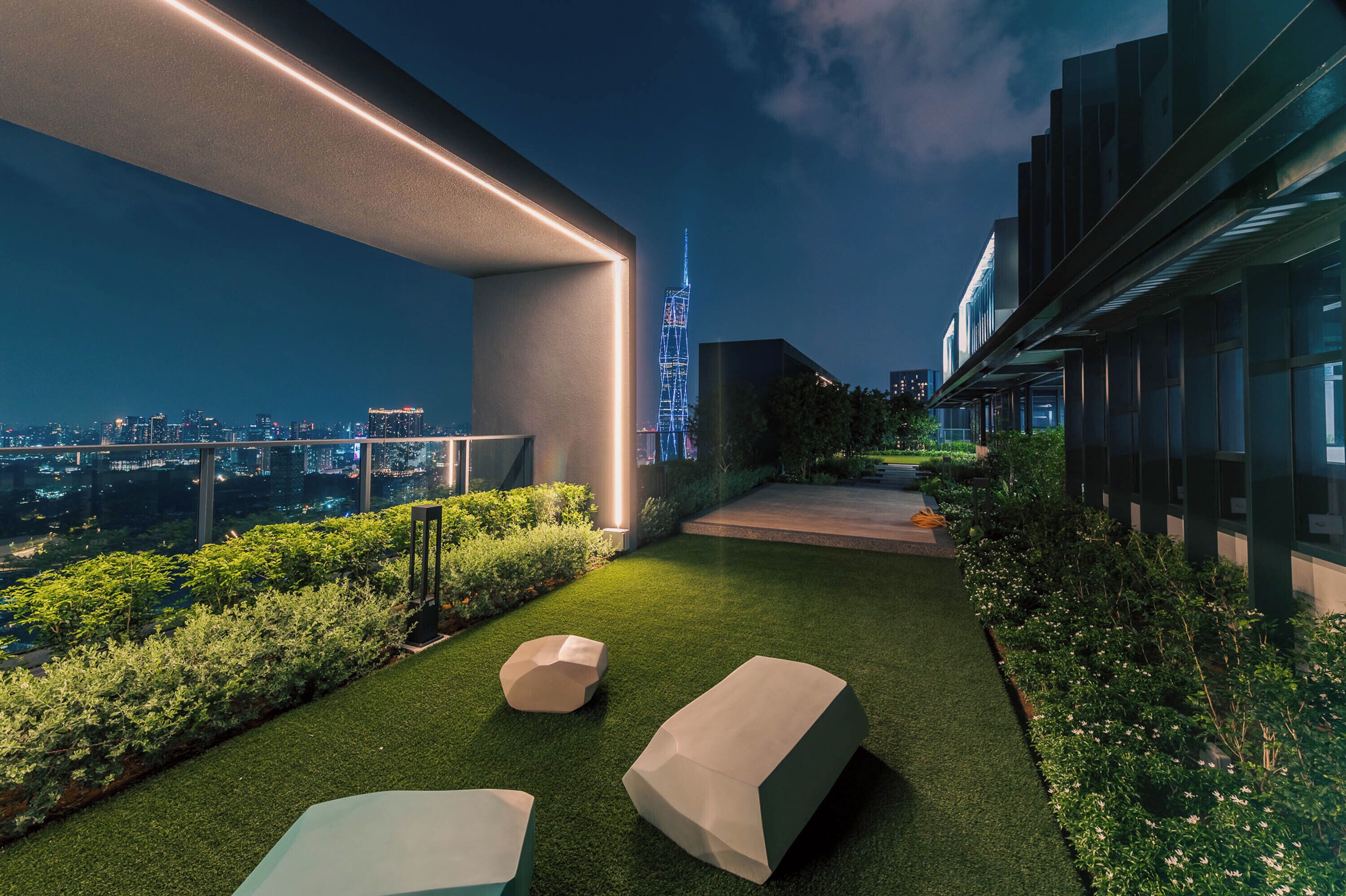 Une terrasse extérieure moderne, dotée d’un revêtement de sol en gazon synthétique, de mobilier aux lignes géométriques et offrant une vue sur les toits illuminés de la ville la nuit.