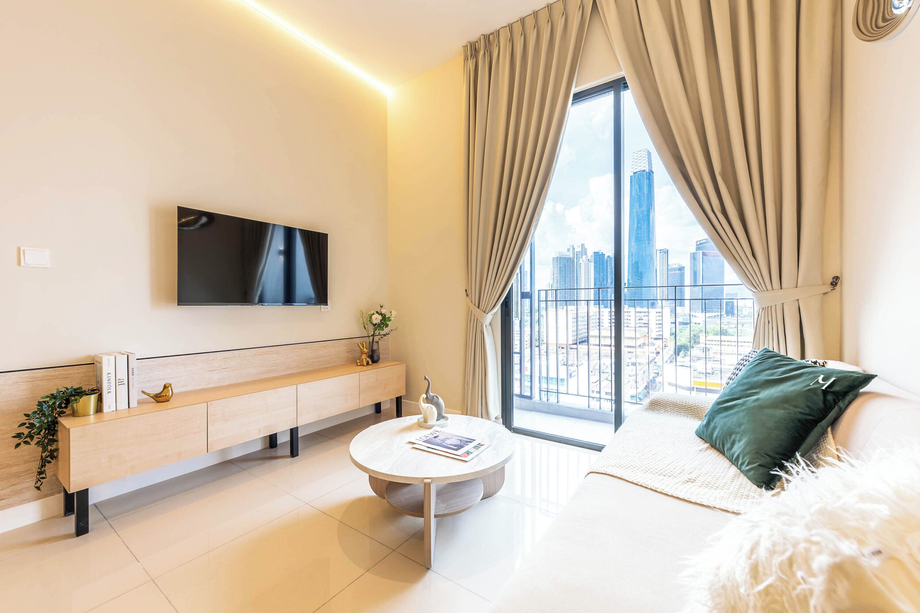 Un salon moderne doté d’un téléviseur à écran plat, d’une table basse ronde, d’un canapé agrémenté d’un coussin vert et offrant une vue sur le paysage urbain grâce à de grandes fenêtres.