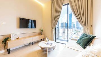 Un salon moderne doté d’un téléviseur à écran plat, d’une table basse ronde, d’un canapé agrémenté d’un coussin vert et offrant une vue sur le paysage urbain grâce à de grandes fenêtres.