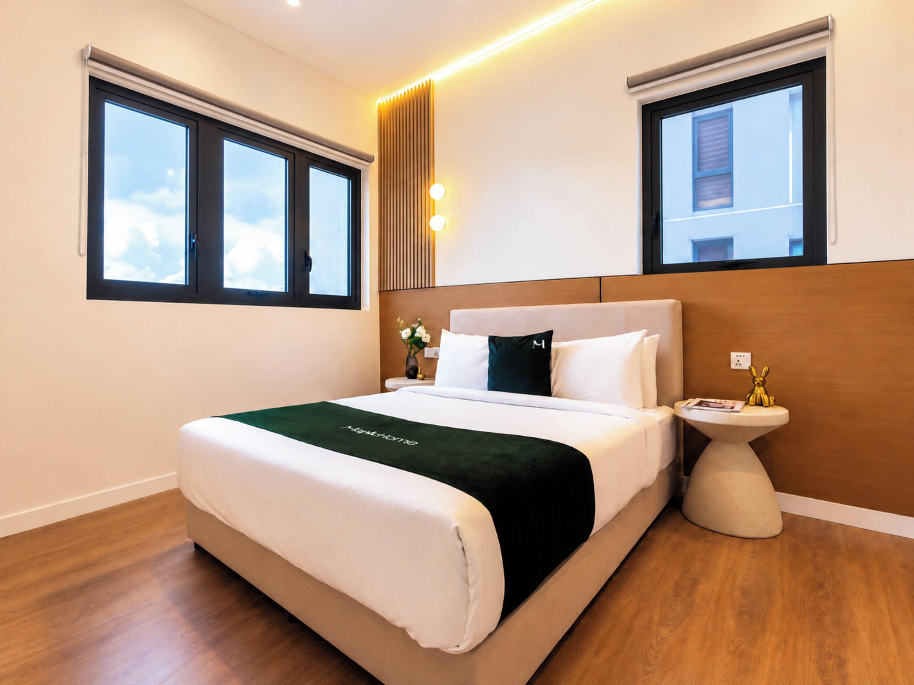 A hotel room with a large bed, two windows, a bedside table with a lamp, and a wall-mounted light fixture.