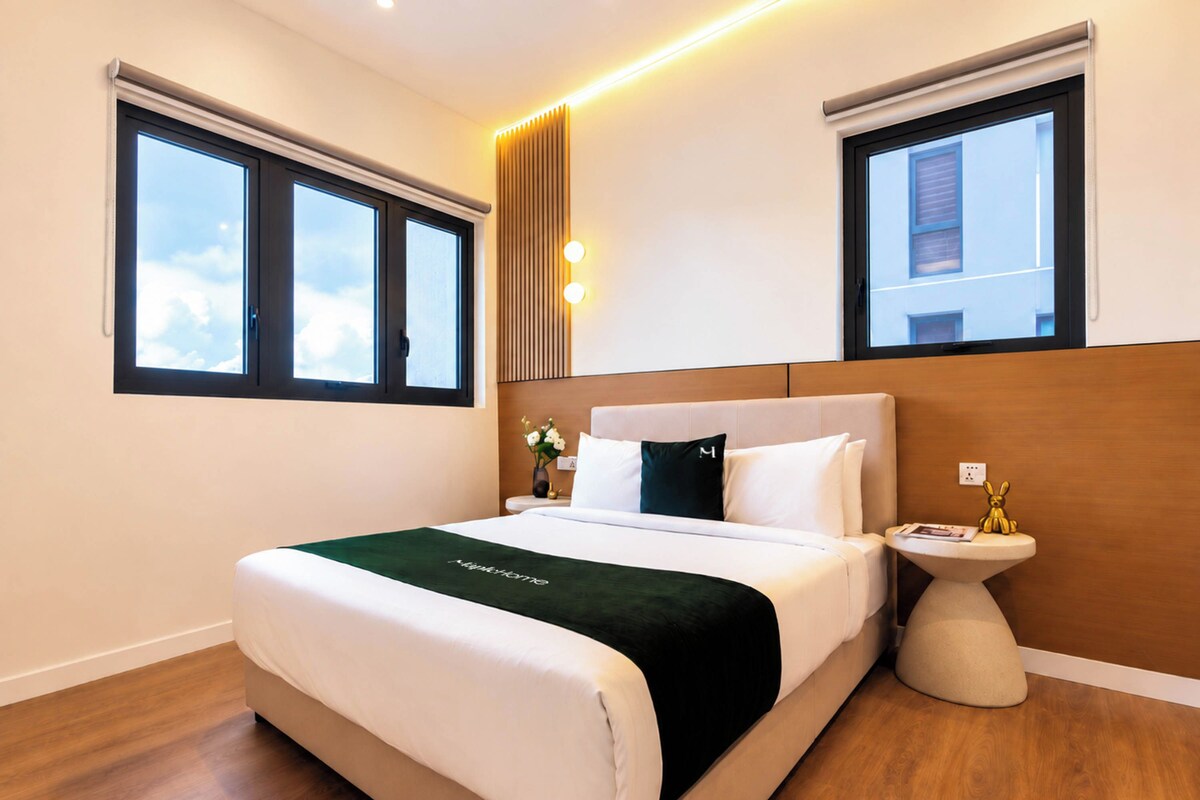 Quarto de hotel com uma cama grande, duas janelas, uma mesa de cabeceira com abajur e luminária na parede.