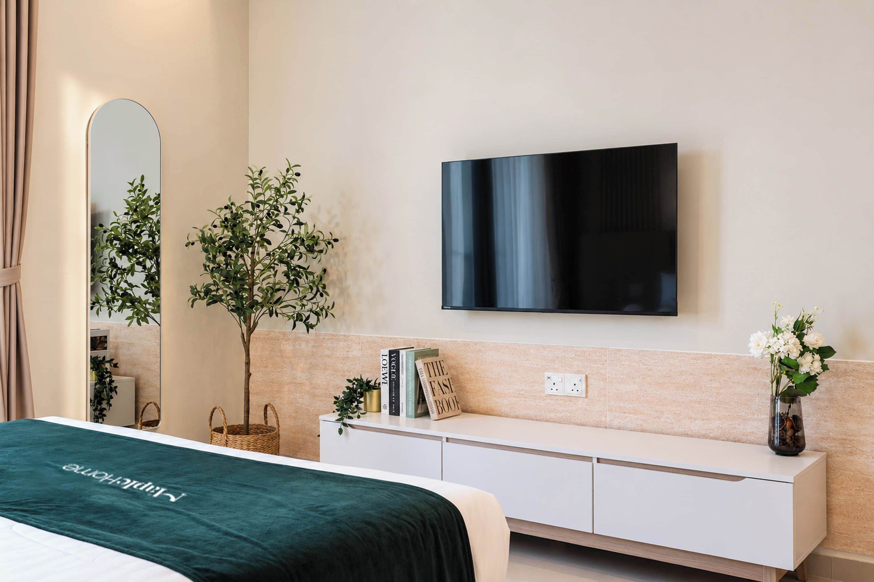 A modern bedroom with a bed, a TV mounted on the wall, a bedside table with books and a sign, and a small plant.
