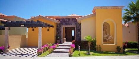 A yellow building with a stone entrance, a covered patio, and a well-maintained garden with pink flowers.