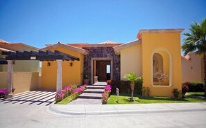 A yellow building with a stone entrance, a covered patio, and a well-maintained garden with pink flowers. - Luxury Montecristo Villa, Private Infinity Pool, Pueblo Bonito Resort (Cabo San Lucas)