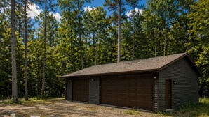 A single-story building with a brown garage door, surrounded by a forested area with tall trees. - Northwoods Lakefront Retreat! Swim, Fish, Relax (Rhinelander)