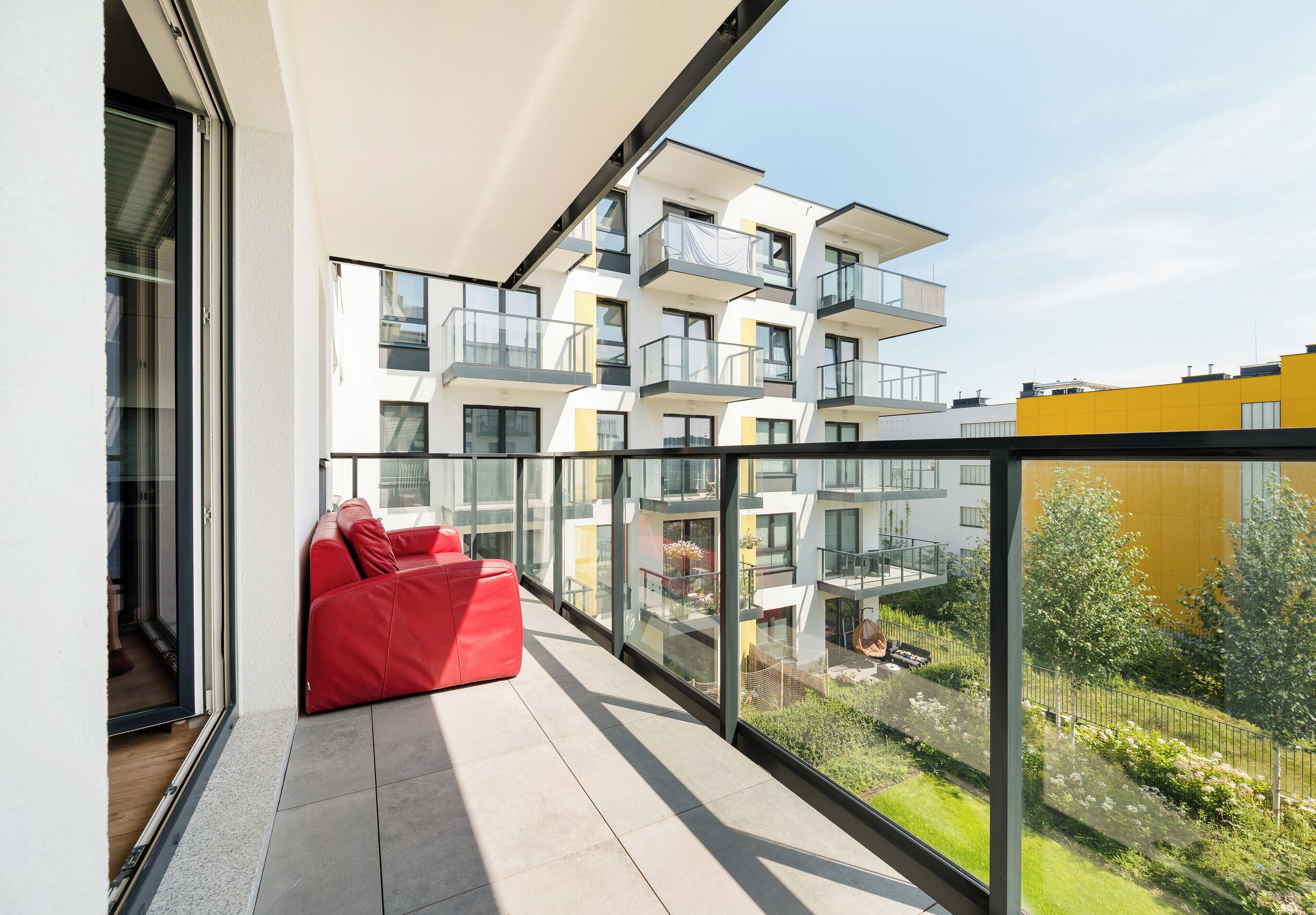 A modern balcony with a red sofa, glass walls, and a view of a multi-story building.