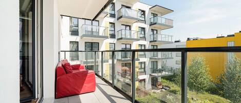 A modern balcony with a red sofa, glass walls, and a view of a multi-story building.