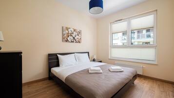 A bedroom with a bed, a nightstand, a lamp, a window with blinds, and a painting on the wall.