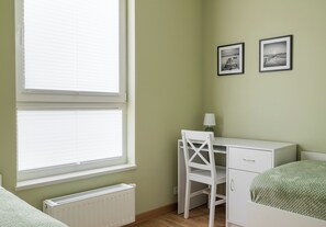 Ein Zimmer mit Fenster, Heizkörper, Schreibtisch, Stuhl, Bett und zwei gerahmten Bildern an der Wand.