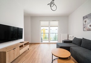 A modern living room with a flat-screen TV, a grey sofa, a wooden coffee table, and a large window with blinds. - Modlińska Riverside Comfort Apartment (Warszawa)