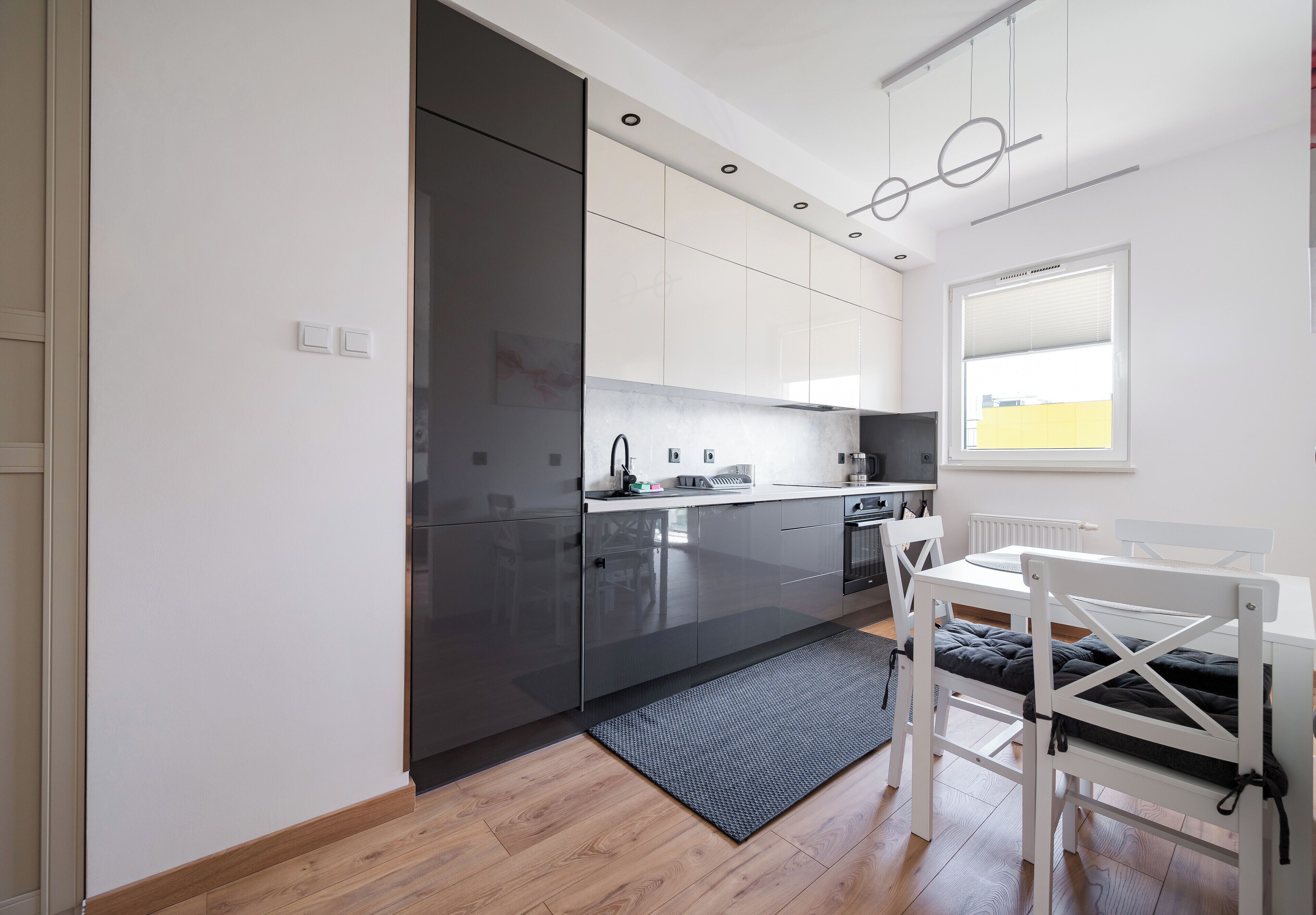 A modern kitchen with a built-in oven, a dining area with a white table and chairs, and a window with blinds.