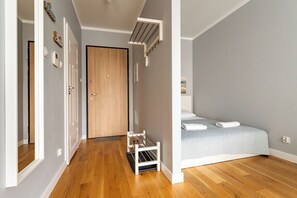 Ein schmaler Flur führt zu einem Schlafzimmer mit einem Bett, einer kleinen weißen Regalwand und einer Holztür.