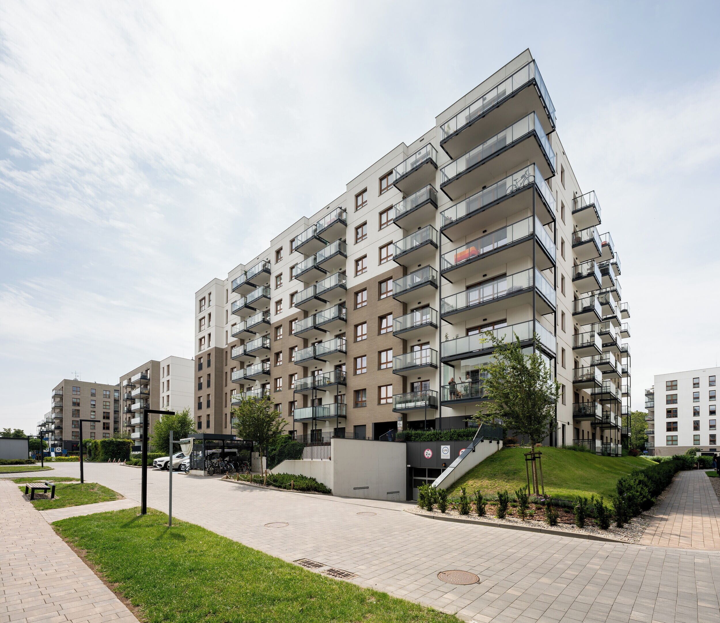 A modern multi-story residential building with balconies, a paved walkway, and a small park area.