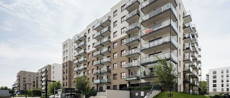 A modern multi-story residential building with balconies, a paved walkway, and a small park area.