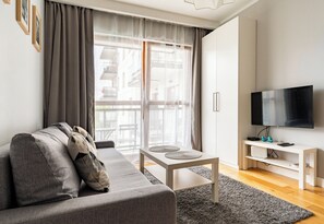 Ein modernes Wohnzimmer mit einem grauen Sofa, einem kleinen weißen Tisch, einem grauen Teppich und einem Fernseher auf einem weißen Schrank.