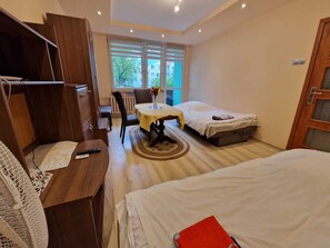 A hotel room with two beds, a wooden wardrobe, a small table with a vase, and a view of the outside through large windows.