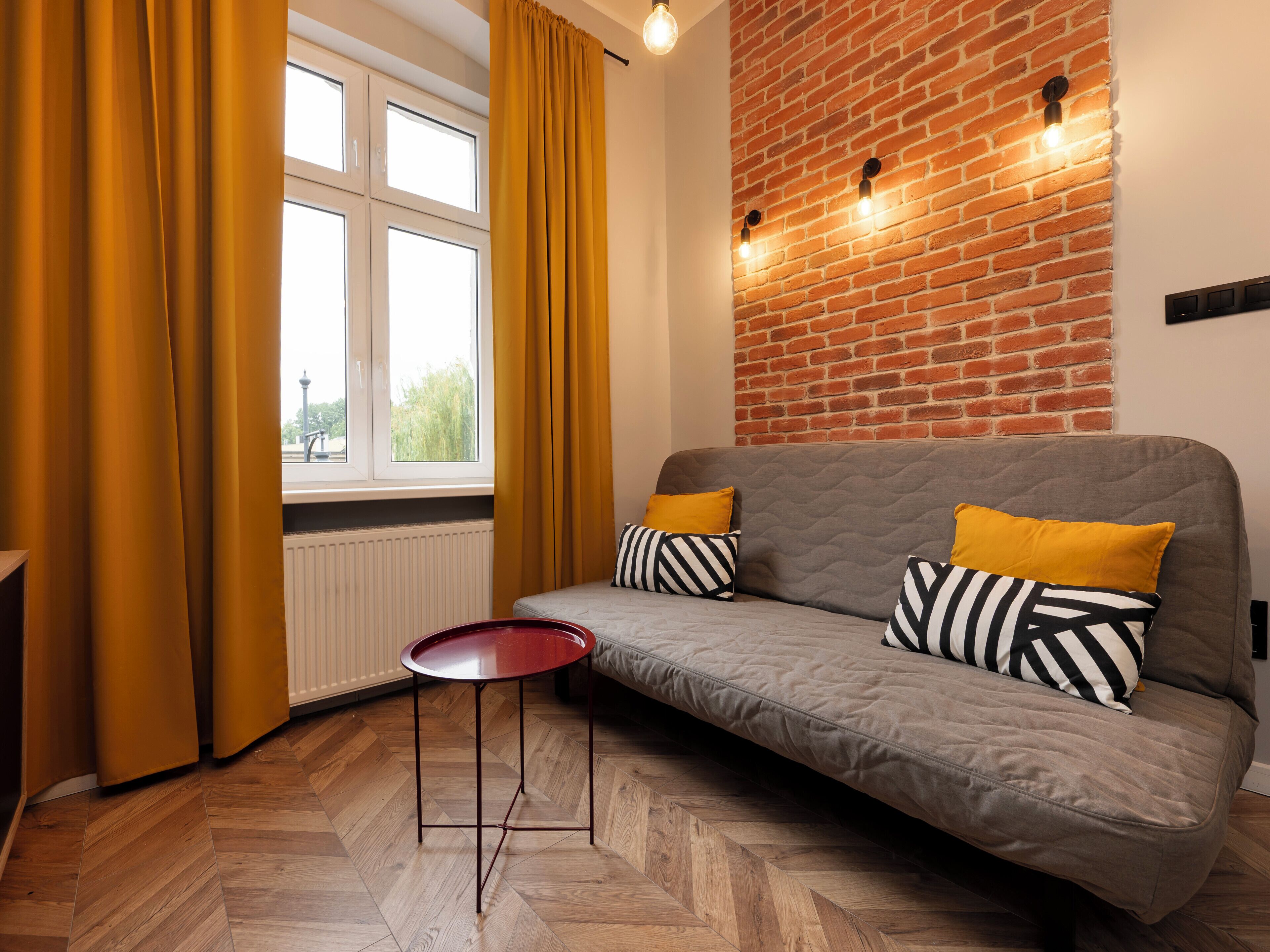 A modern living room with a sofa, a small table, a window with curtains, a brick wall, and a radiator.